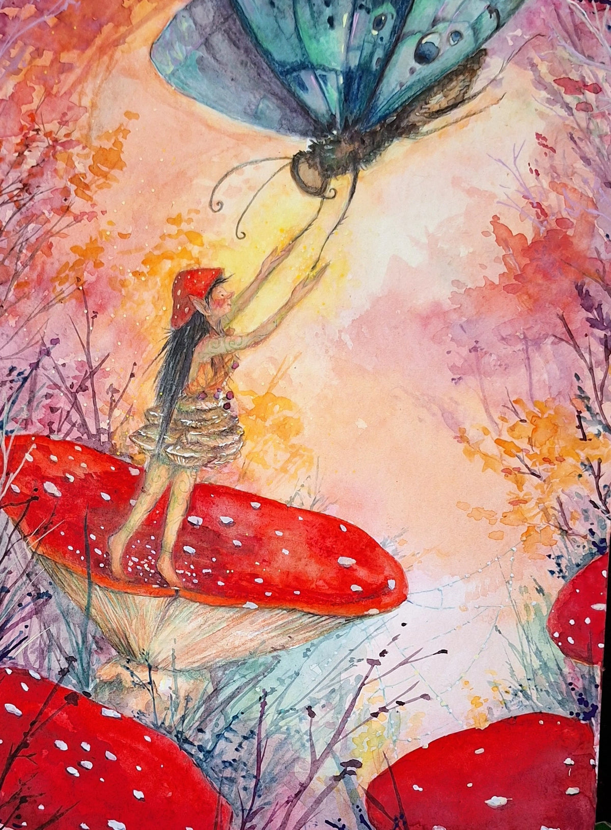 Postkarte Herbstwesen von Anne Gies A6: ein kleines Wesen mit roter Mütze steht auf einem Fliegenpilz und hält sich an einem großen blaugrauen Schmetterling fest, ringsum leuchtendes Herbstlaub