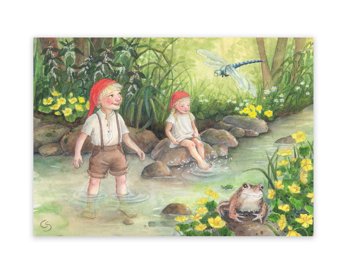 Postkarte Am Bach von Corinna Schwerdtfeger A6: zwei Wichtelkinder spielen barfuß im Bach zwischen gelben Sumpfdotterblumen, eine Libelle und ein Frosch schauen zu