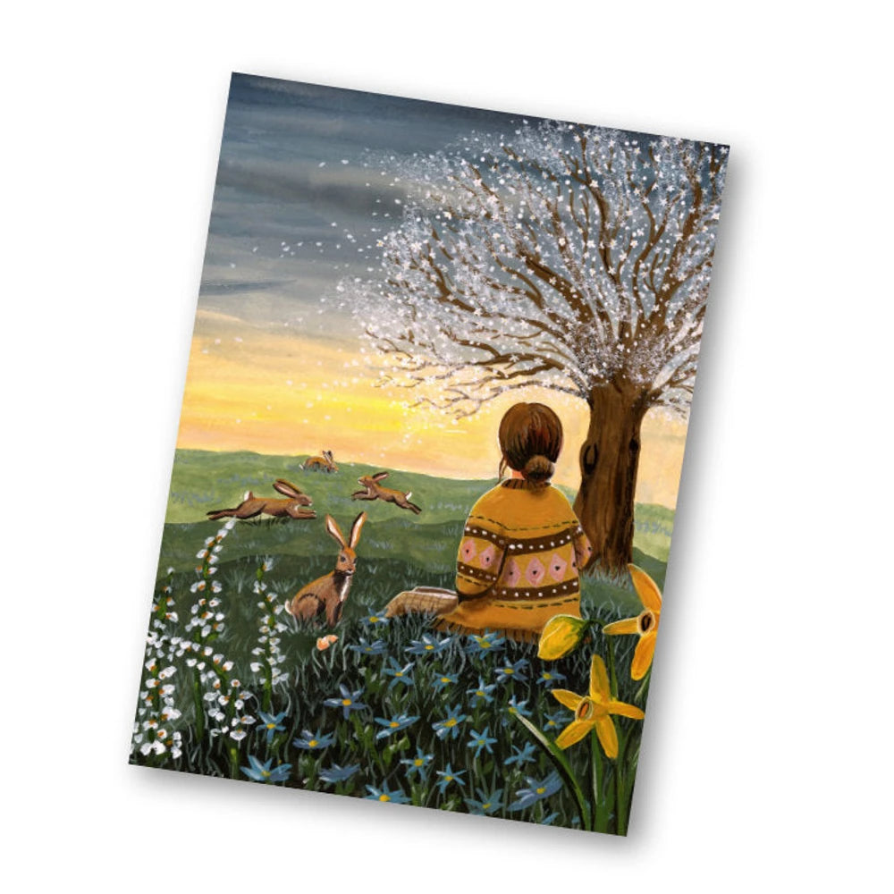 Postkarte Osterfeld von Esther Bennink A6: ein Mädchen sitzt bei Sonnenaufgang auf einer Frühlingswiese unter einem Baum, Hasen und Narzissen ringsum