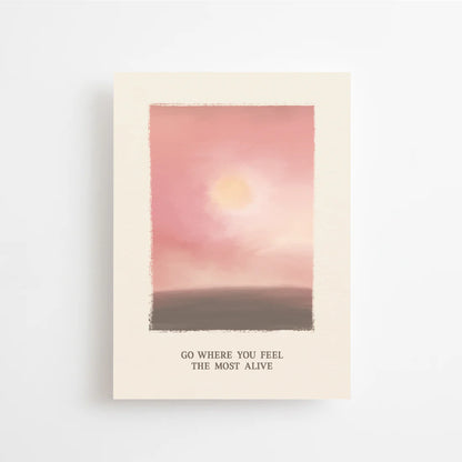 Postkarte Go where you feel the most alive von Anna Cosma A6: eine Karte mit gemaltem Abendhimmel in Rosé und Rot, einer tief stehenden Sonne über dunklem Land und dem Schriftzug „Go where you feel the most alive“