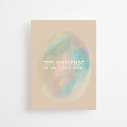Postkarte The universe is on your side von Anna Cosma A6: eine beige Karte mit einem pastellfarbenen, schimmernden Oval wie ein Nebel im All und dem Schriftzug „The universe is on your side“