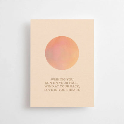 Postkarte Wishing you sun on your face, wind at your back, love in your heart von Anna Cosma A6: eine pfirsichfarbene Karte mit einer Sonne in Orange und Rosé und dem Schriftzug „Wishing you sun on your face, wind at your back, love in your heart“