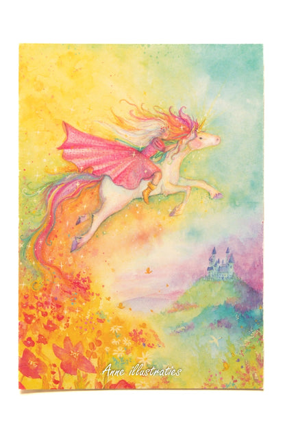 Postkarte Einhorn von Anne Gies A6: eine Gestalt im rosa Umhang reitet auf einem weißen Einhorn mit Regenbogenmähne durch einen bunten Himmel, unten Blumen und ein kleines Schloss