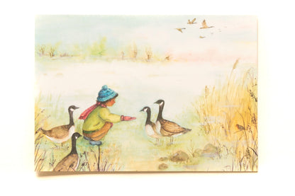 Postkarte Gänse von Anne Gies A6: ein Kind mit blauer Mütze und rotem Schal hockt am Seeufer und füttert Wildgänse, am Himmel ziehen weitere Gänse vorbei