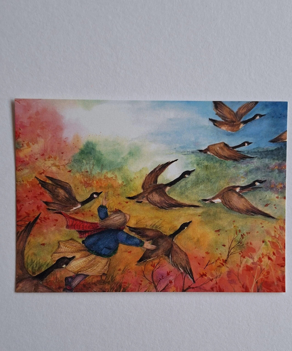 Postkarte Gänseflug von Anne Gies A6: ein Kind in blauer Jacke mit rotem Schal fliegt mit einer Schar Wildgänse über eine bunte Herbstlandschaft