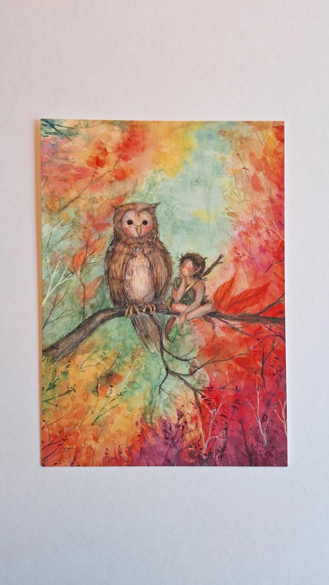 Postkarte Herbst-Eule von Anne Gies A6: eine braune Eule sitzt auf einem Ast neben einem kleinen Wesen in Grün, ringsum leuchtendes Herbstlaub