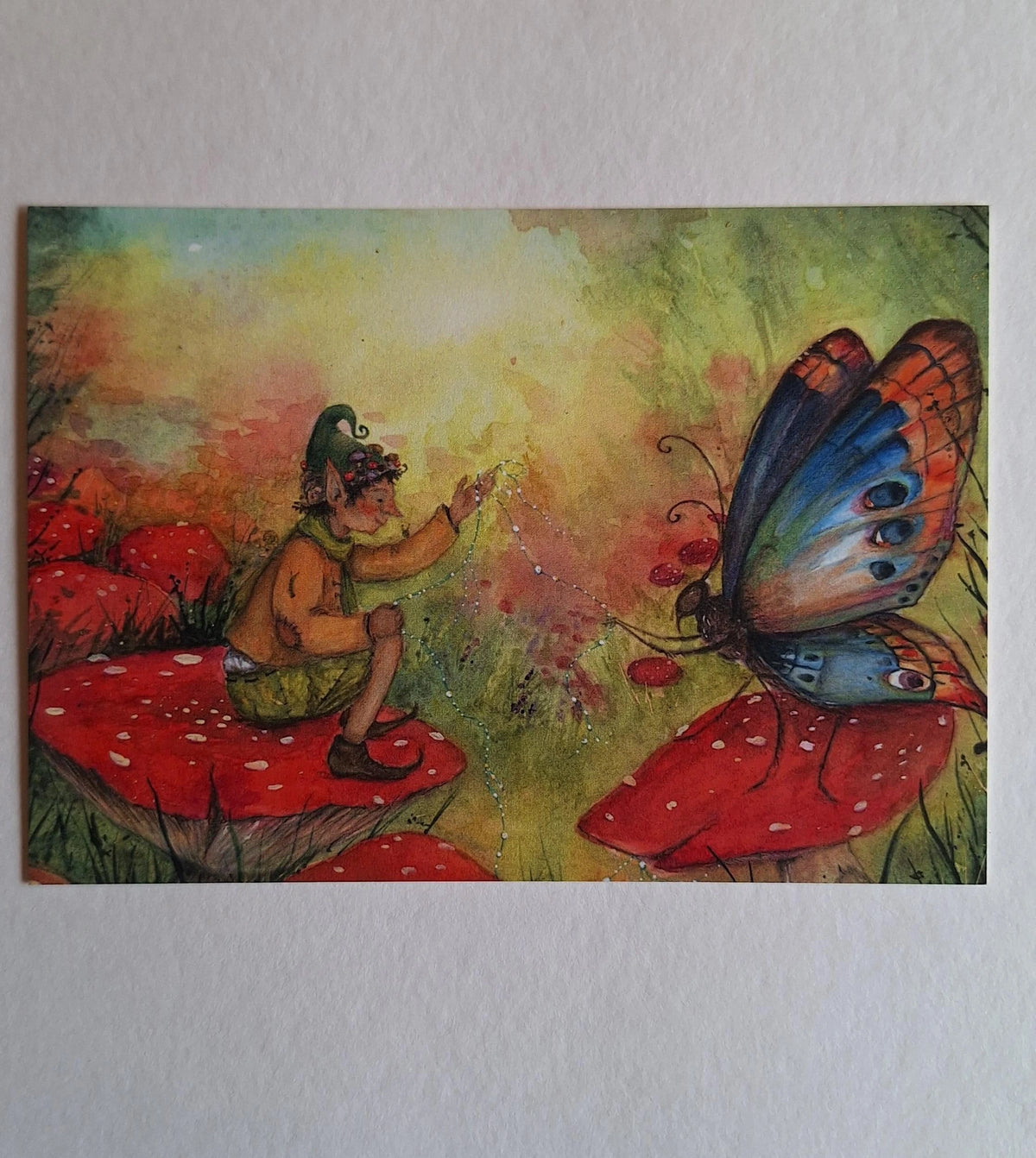 Postkarte Herbsttraum von Anne Gies A6: ein Wichtel mit grüner Zipfelmütze sitzt auf einem Fliegenpilz und streckt die Hand nach einem großen bunten Schmetterling aus