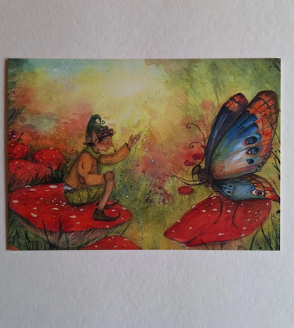 Postkarte Herbsttraum von Anne Gies A6: ein Wichtel mit grüner Zipfelmütze sitzt auf einem Fliegenpilz und streckt die Hand nach einem großen bunten Schmetterling aus