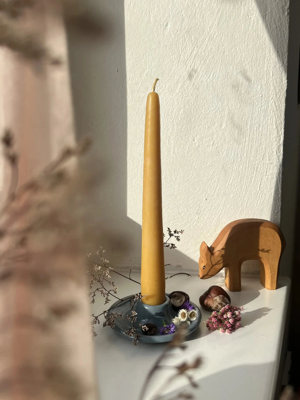 Kerzenhalter aus Keramik graublau von El Puente mit gelber Stabkerze am Fenster, daneben ein Holzreh, Foto