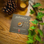 Emaille-Pin Schnecke patience von Esther Bennink, Emaille-Pin Schnecke auf einer dunklen Karte mit dem Spruch patience