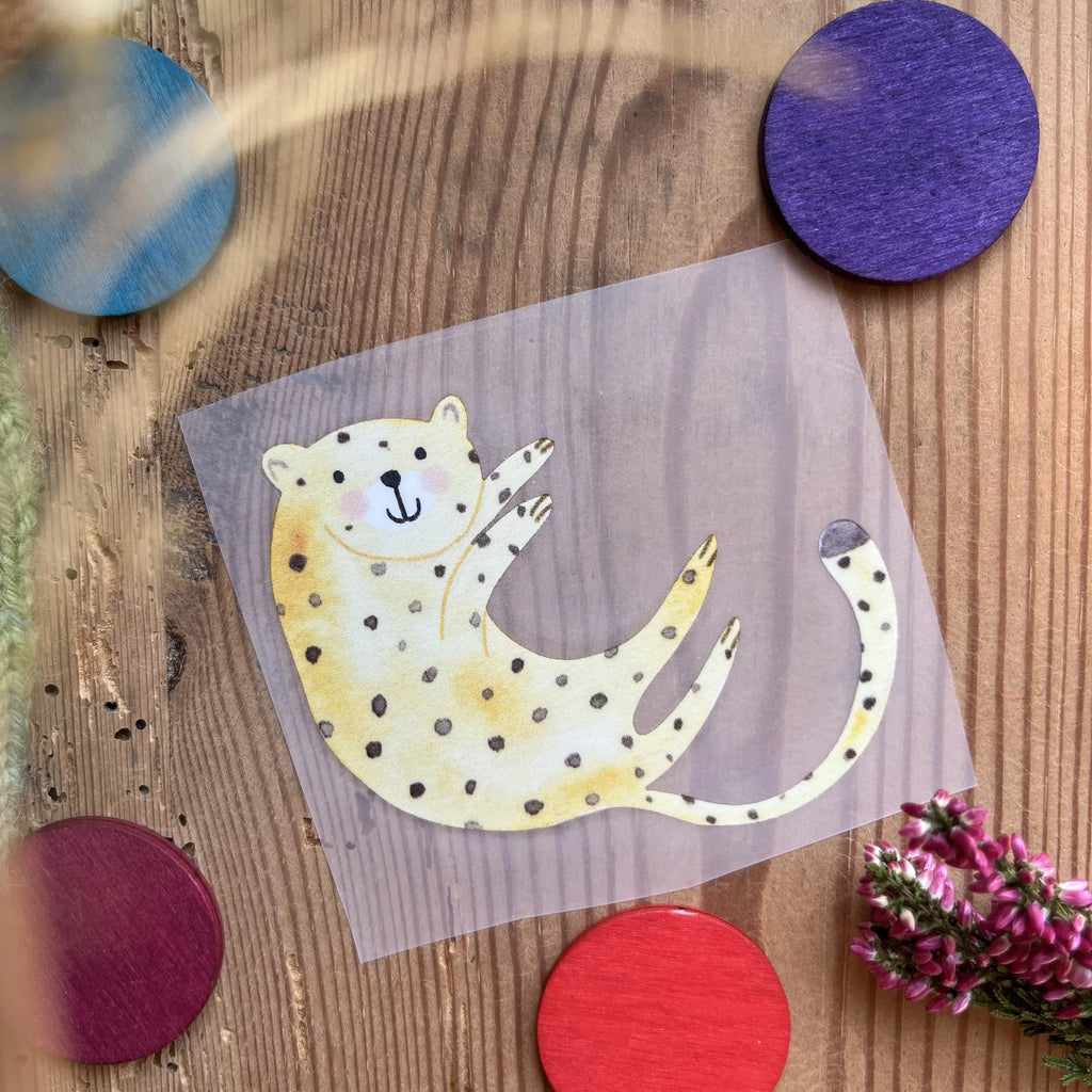 Hannahs Firlefanz Bügelbild verspielter Gepard auf dem Rücken, auf Holz zwischen bunten Holzscheiben