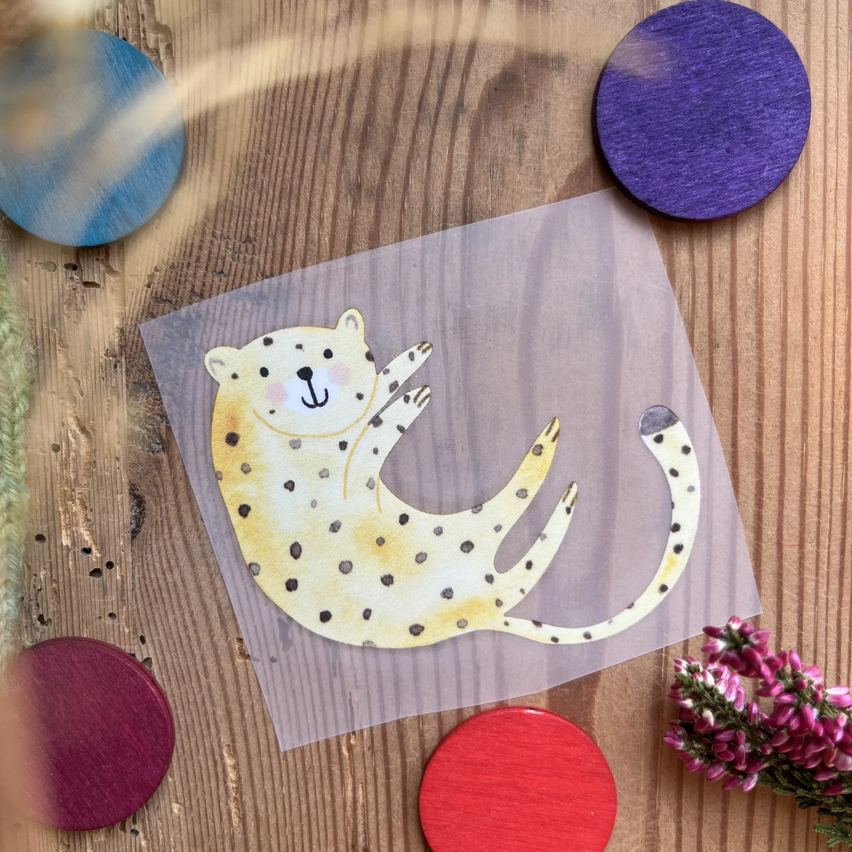 Hannahs Firlefanz Bügelbild verspielter Gepard auf dem Rücken, auf Holz zwischen bunten Holzscheiben
