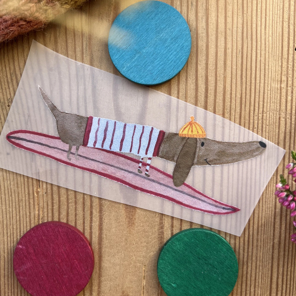 Hannahs Firlefanz Bügelbild Dackel im Ringelpulli auf dem Surfbrett, auf Holz zwischen bunten Holzscheiben