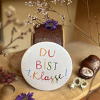 Hannahs Firlefanz Button Du bist 1. Klasse! in bunter Schrift auf Holz zwischen Kastanien und Astern