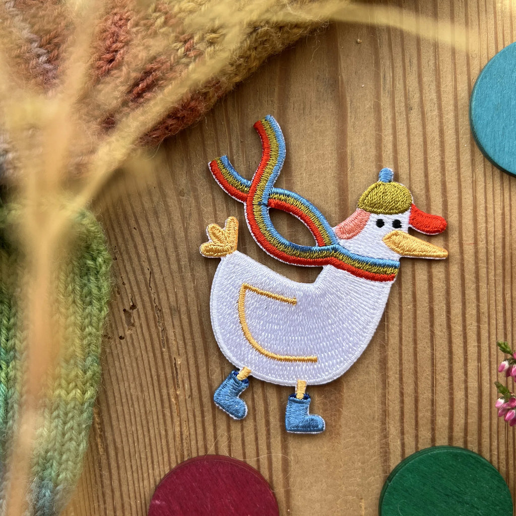 Hannahs Firlefanz Patch Gans mit Regenbogenschal und Mütze, gestickt, auf Holz zwischen bunten Holzscheiben
