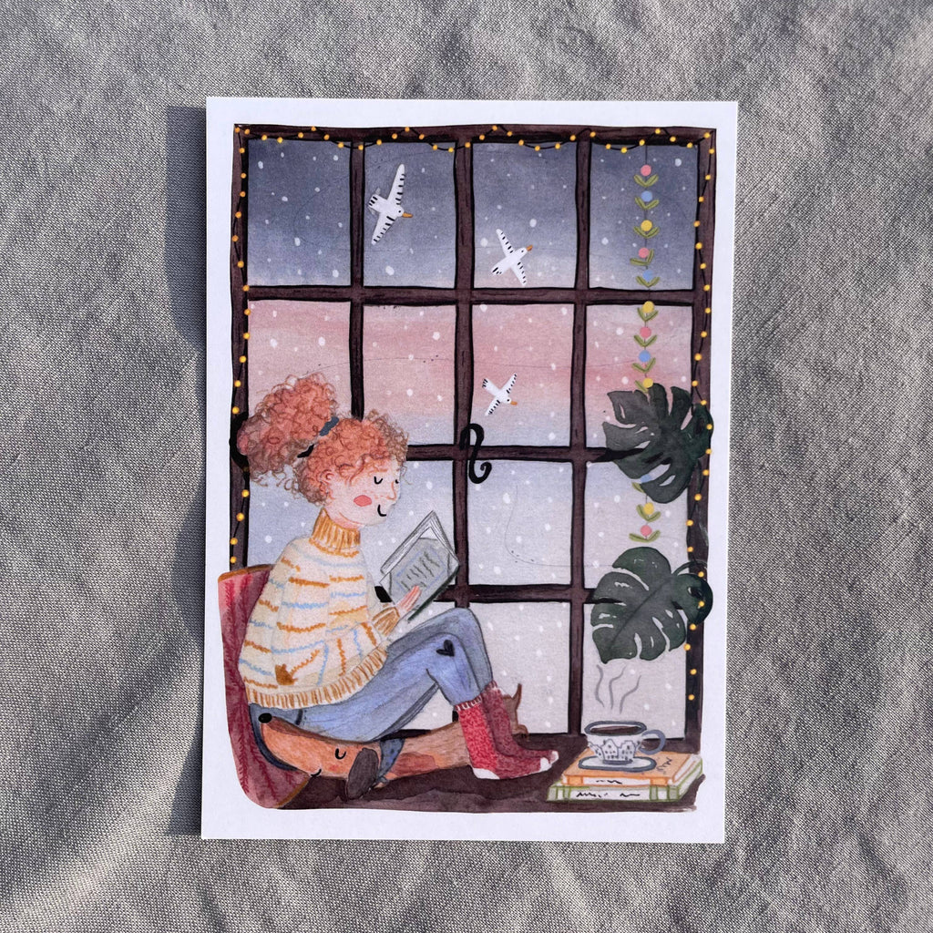 Postkarte Leseabend von Hannahs Firlefanz A6, Person mit roten Locken liest am Fenster, draußen fällt Schnee, daneben Tee und Bücher