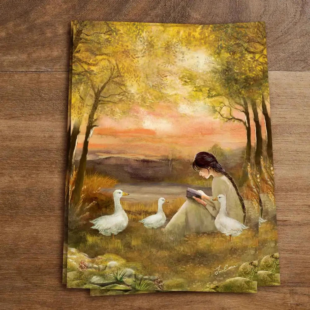 Postkarte Geschichten am See von Iris Esther A6: eine junge Frau liegt mit einem Buch im Gras am Wasser, weiße Gänse spazieren um sie herum