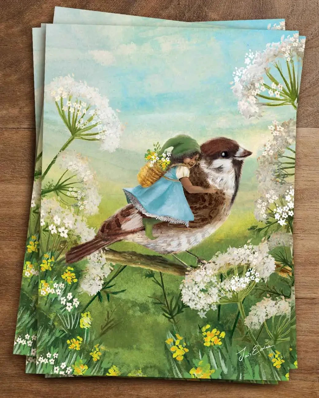 Postkarte Winzige Reise von Iris Esther A6: ein kleines Wichtelmädchen mit grüner Zipfelmütze reitet auf einem Spatz über blühende Wiesen