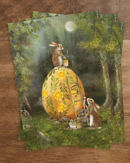 Postkarte Zusammenarbeit an Ostern von Iris Esther A6: zwei Hasen bemalen unter dem Frühlingsmond ein großes goldenes Ei mit Ringelblumen und Ranken