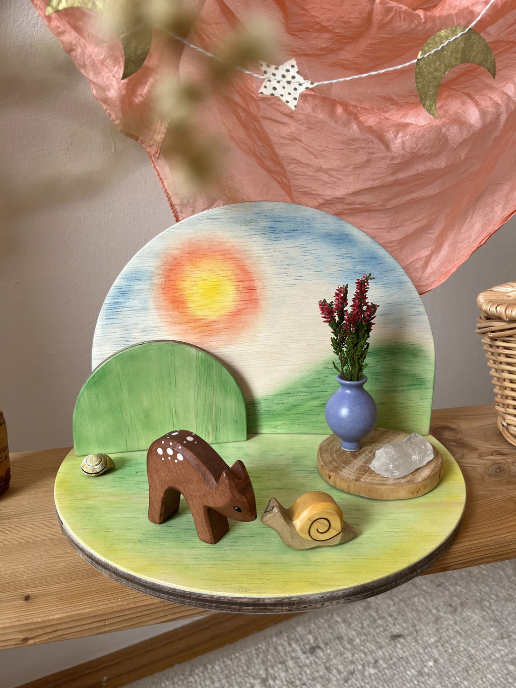 Halbrunder Jahreszeitentisch aus Holz zum Schrauben, bemaltes Beispiel mit Sonne am Himmel, grünem Hügel und grüner Bodenplatte, dekoriert mit Reh, Schnecke, Vase und Bergkristall, Foto