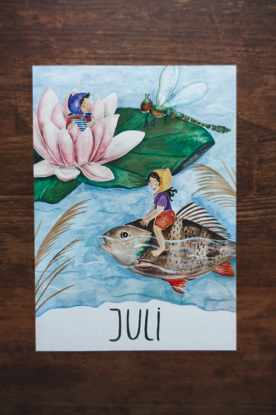 Monatskarte Juli aus dem Set von Joringl, Wichtel reitet auf einem Fisch, Seerose und Libelle, auf Holz fotografiert