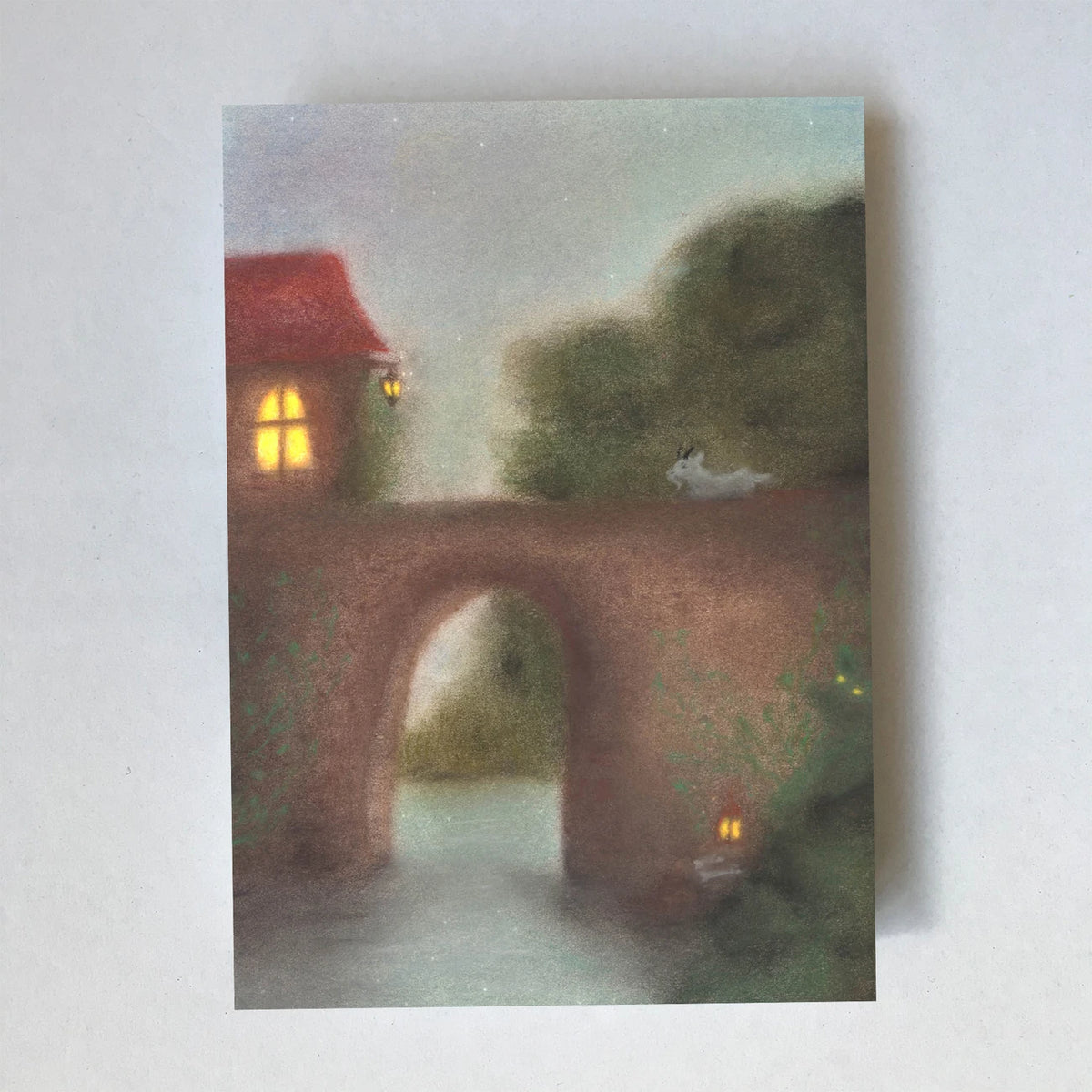 Postkarte Brücke von Lot Bouwes A6: steinerne Bogenbrücke über einen Fluss, ein rotes Haus mit Licht, ein kleines Tier auf der Brücke, kleines Licht am Ufer
