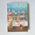 Postkarte Fenster von Lot Bouwes A6: Blick durch ein Sprossenfenster auf ein verschneites Dorf mit warm leuchtenden Häusern, auf der Fensterbank Katze und eine Kerze