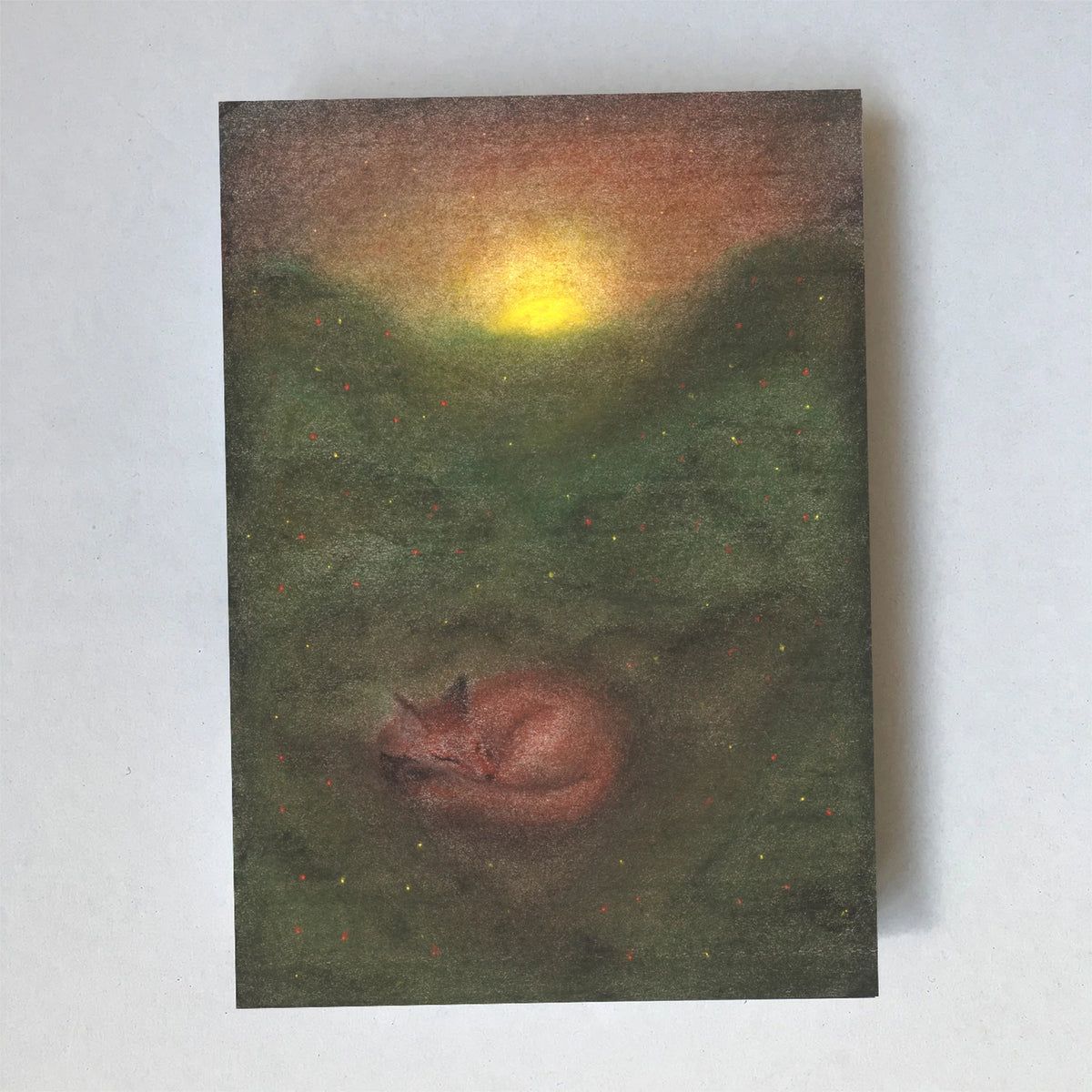 Postkarte Fuchs von Lot Bouwes A6: ein Fuchs schläft eingerollt auf einer grünen Lichtung, die Sonne geht hinter dem Wald unter