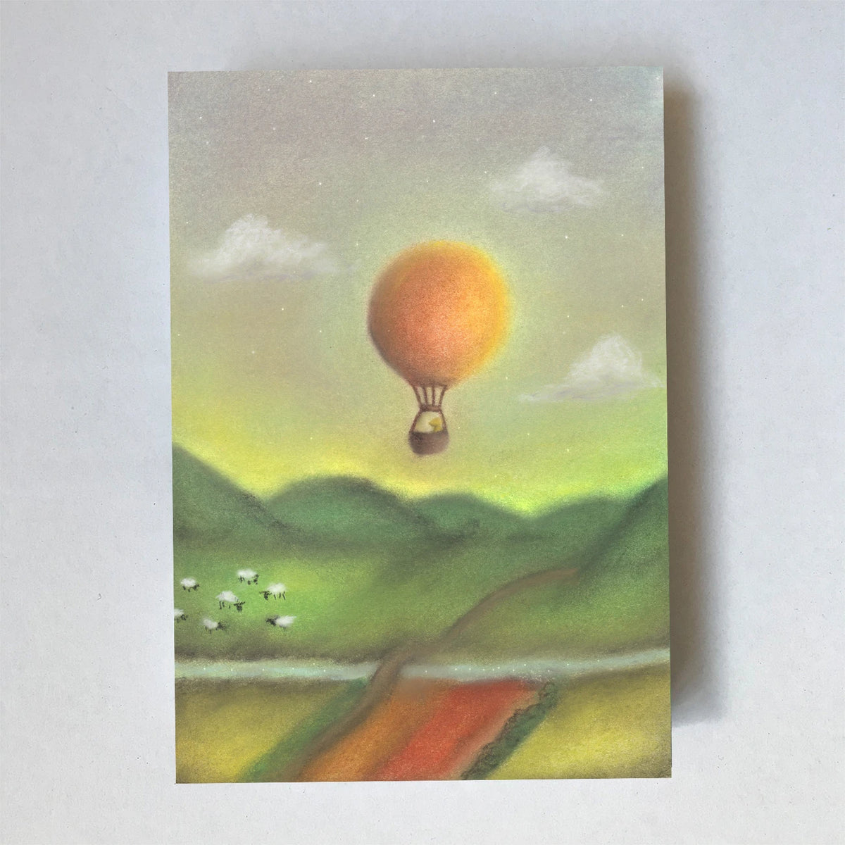 Postkarte Heißluftballon von Lot Bouwes A6: orangefarbener Heißluftballon über grünen Hügeln, Schafe auf der Wiese, Felder, Wolken