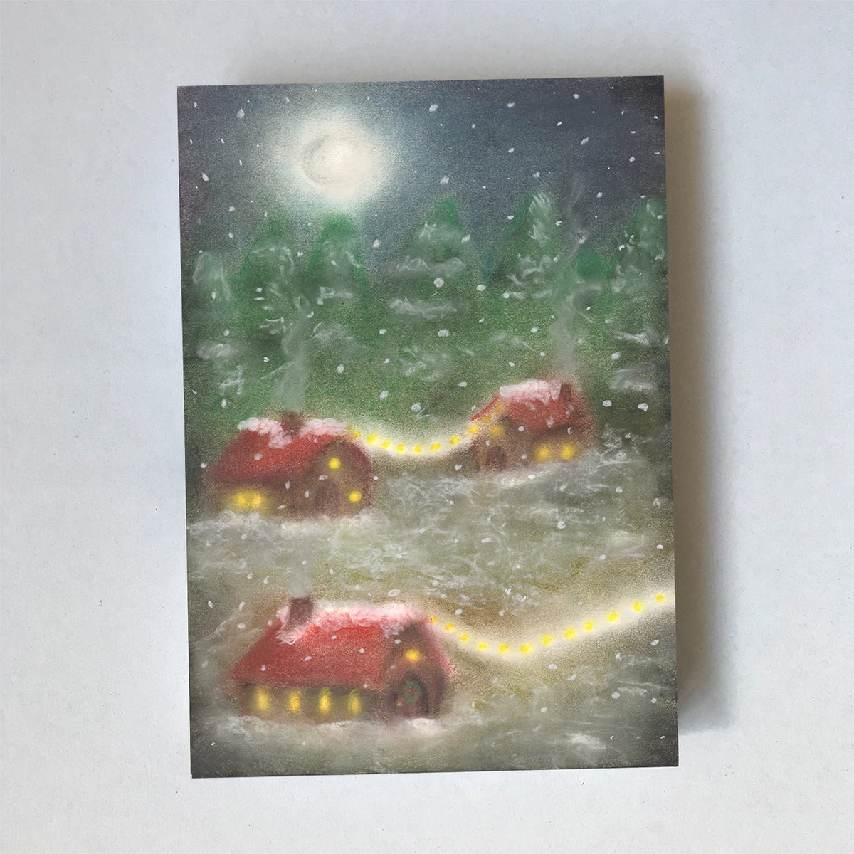Postkarte Winterdorf von Lot Bouwes A6: verschneites Dorf mit roten Häuschen bei Nacht, Lichterketten führen von Haus zu Haus, Vollmond, Tannen, Schneeflocken