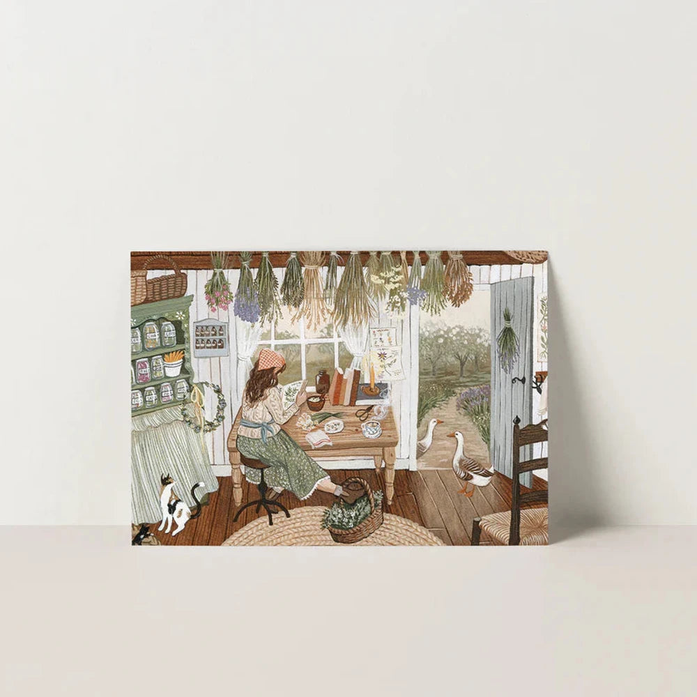 Postkarte Die Kräuterkundige von Robin Pieterse A6: eine Frau sitzt zwischen getrockneten Kräuterbündeln an ihrem Tisch und studiert botanische Notizen, durch die offene Tür sieht man den Garten mit Gänsen