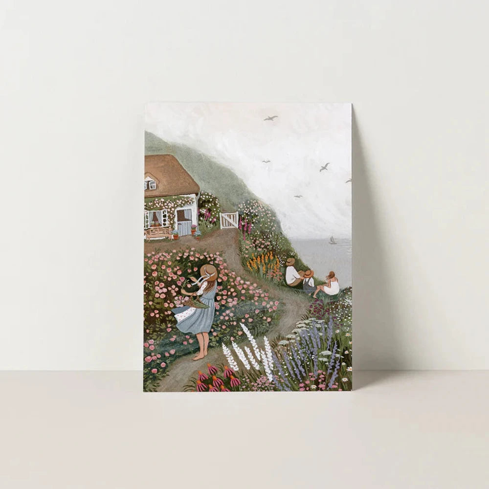 Postkarte Rosenhäuschen von Robin Pieterse A6: ein Häuschen voller Kletterrosen über dem Meer, eine Familie genießt den Nachmittag, eine Frau pflückt Blumen