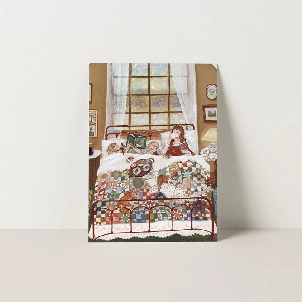 Postkarte Winternest von Robin Pieterse A6: eine Familie liegt unter einer Patchworkdecke im Bett, liest und frühstückt, eine Katze schläft am Fußende