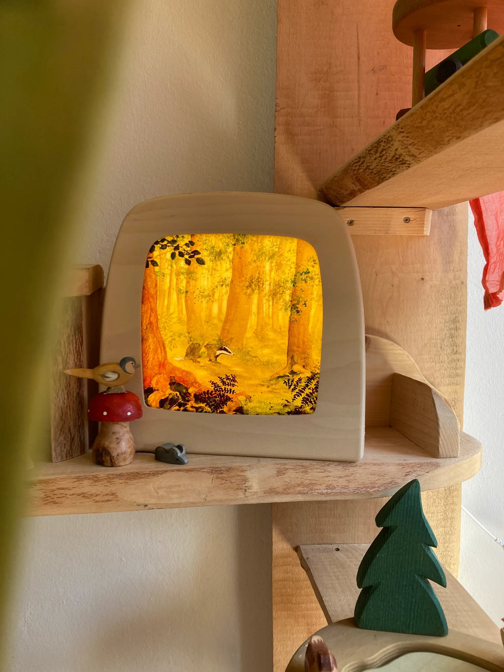 Magische Lampe Toverlux mit leuchtendem goldenen Herbstwald auf einem Wandregal neben Pilzfigur und Holztanne
